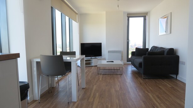 A modern living room with a flat-screen TV, a sofa, a dining table, and a chair. - Quay Apartments (Salford)