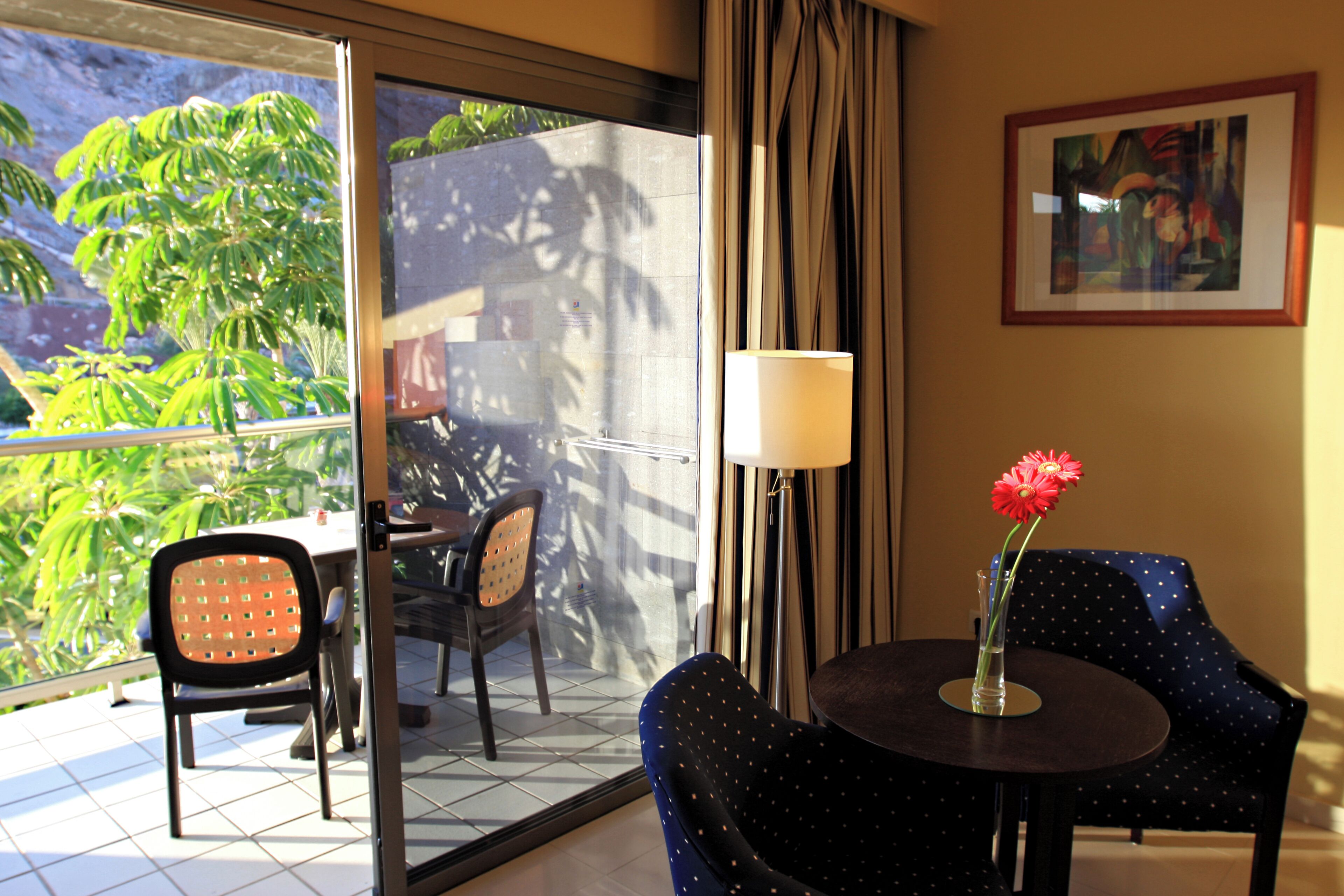 Ein Hotelzimmer mit Blick auf Palmen, einem Essbereich mit Stühlen, einem Tisch mit Blumenvase und einem Gemälde an der Wand.
