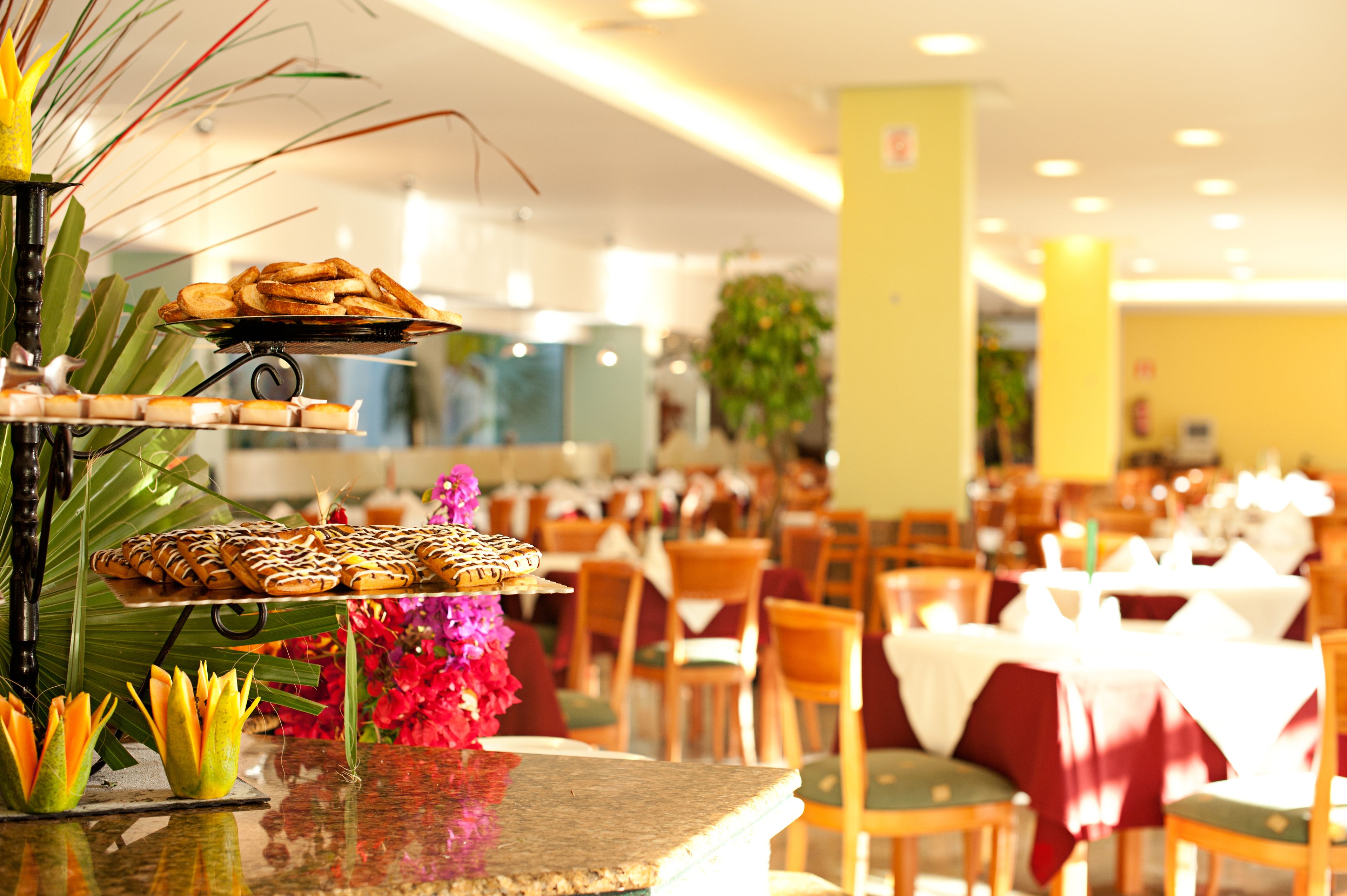 A restaurant with multiple dining tables and chairs, a buffet with various food items, and decorative plants.