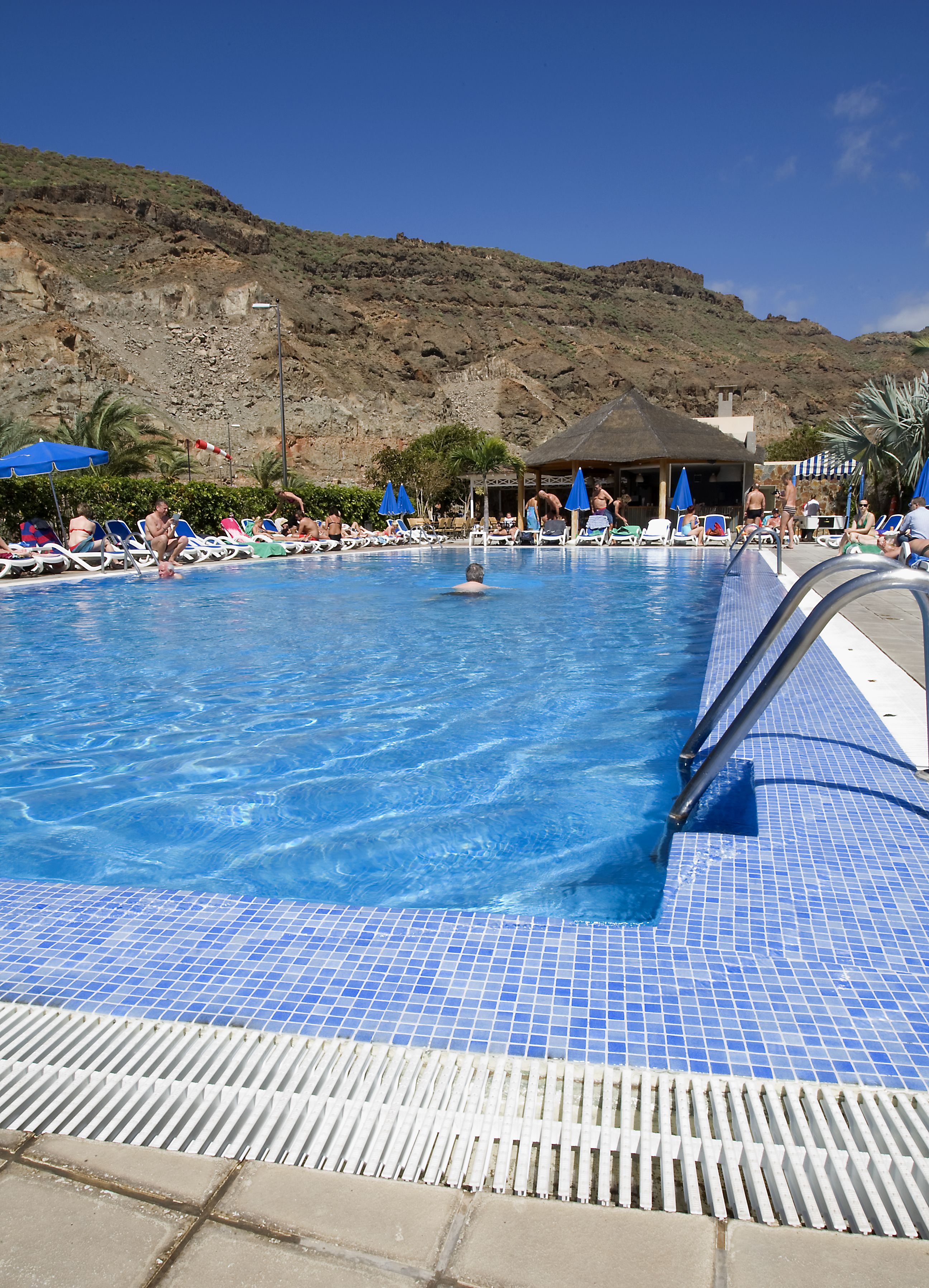 Une piscine à l’eau d’un bleu cristallin, entourée de chaises longues et de parasols, avec en toile de fond un paysage montagneux.