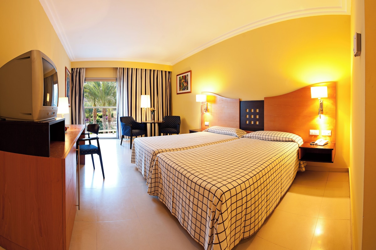 A hotel room with two beds, a desk with a television, a chair, and a window with curtains.