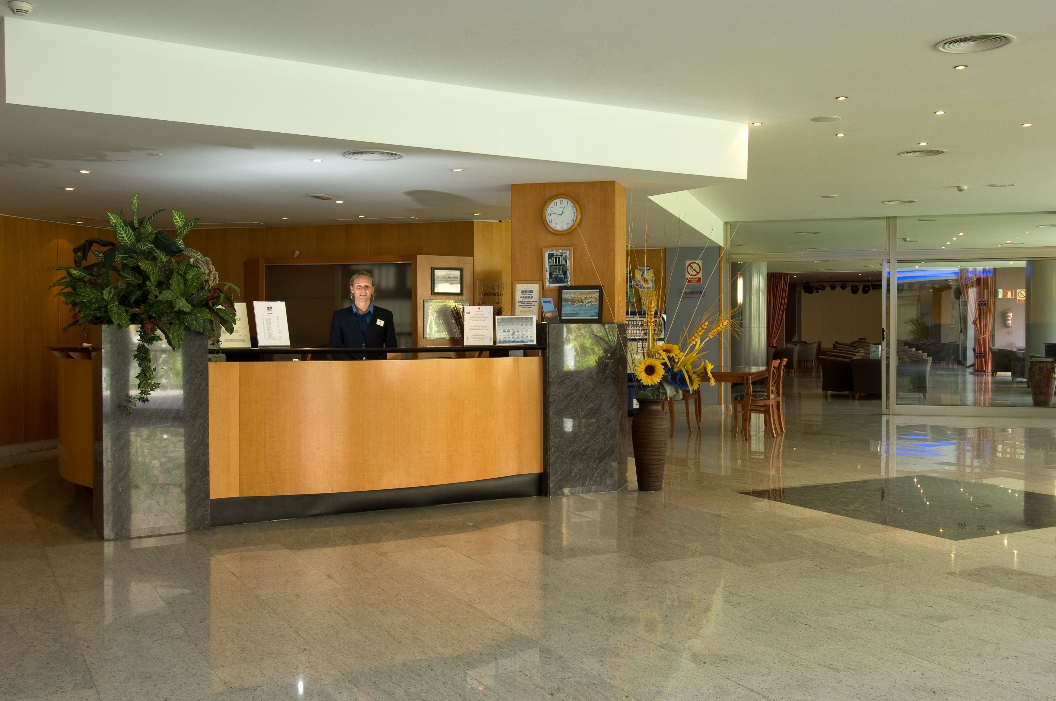 Een hotel receptie met een houten balie, een klok, ingelijste foto’s en een grote potplant.