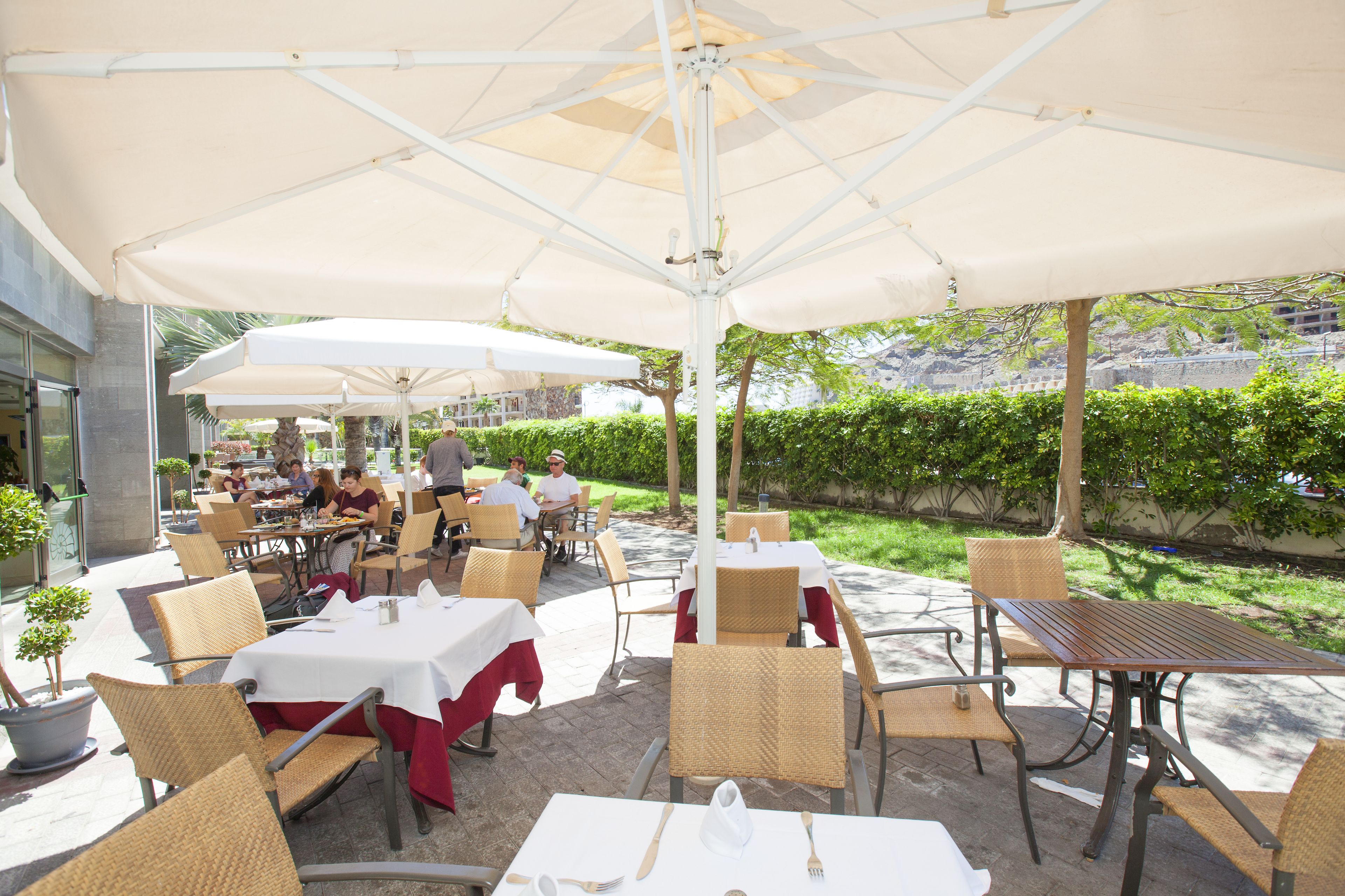 An outdoor patio area with tables and chairs, covered by umbrellas, and surrounded by greenery.