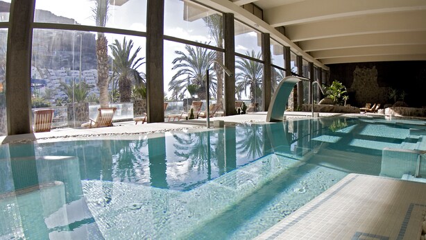 An indoor swimming pool with clear water, large windows, and a view of palm trees and a rocky landscape. - Hotel LIVVO Costa Taurito & Aquapark - All Inclusive (Mogan)