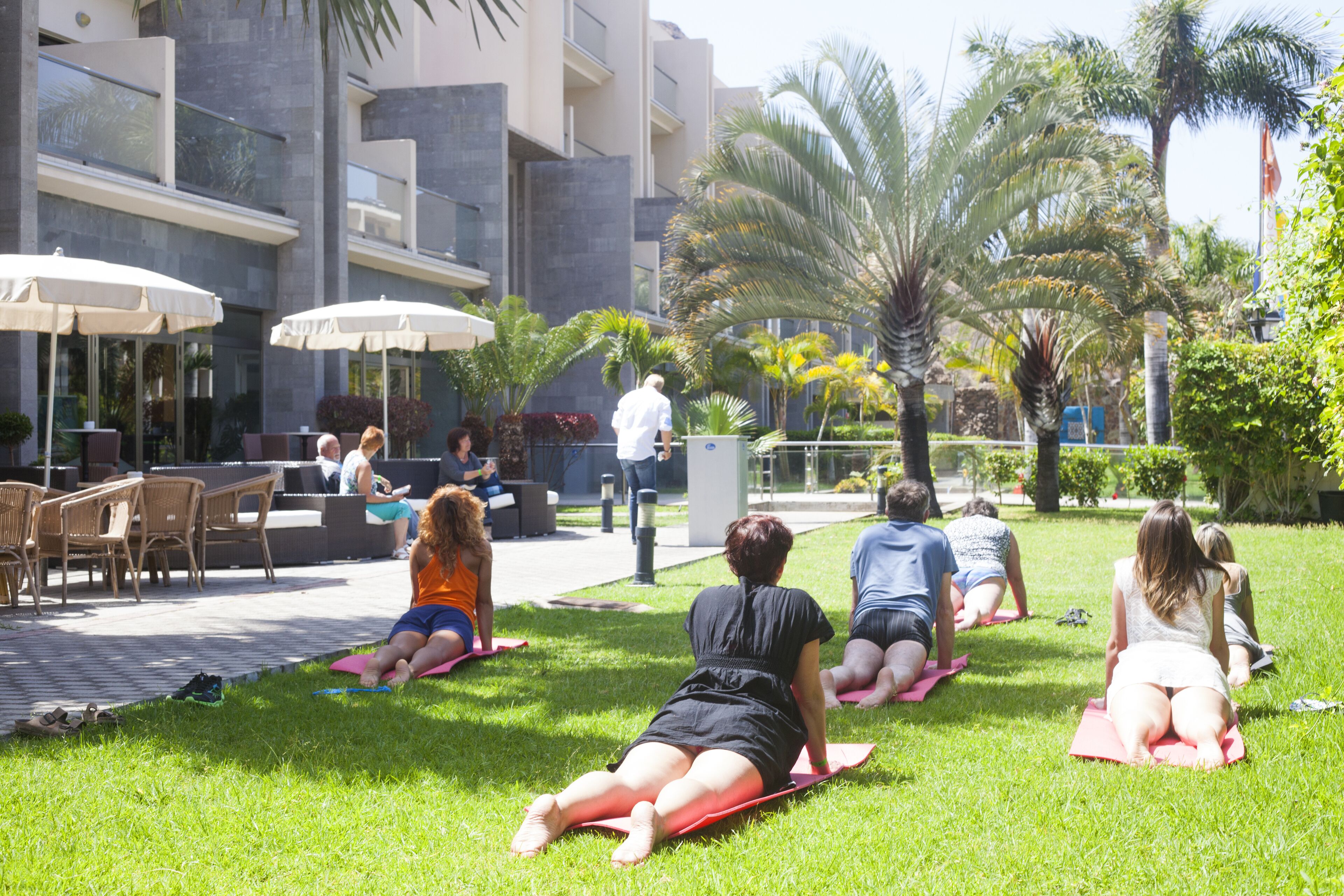 Un groupe de personnes pratiquant le yoga sur l’herbe, dans un espace extérieur aménagé avec des chaises et des parasols.