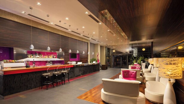 A modern bar area with a counter, bar stools, and a seating area with white sofas and a lamp. - Golden Tulip Ana Dome (Cluj-Napoca)