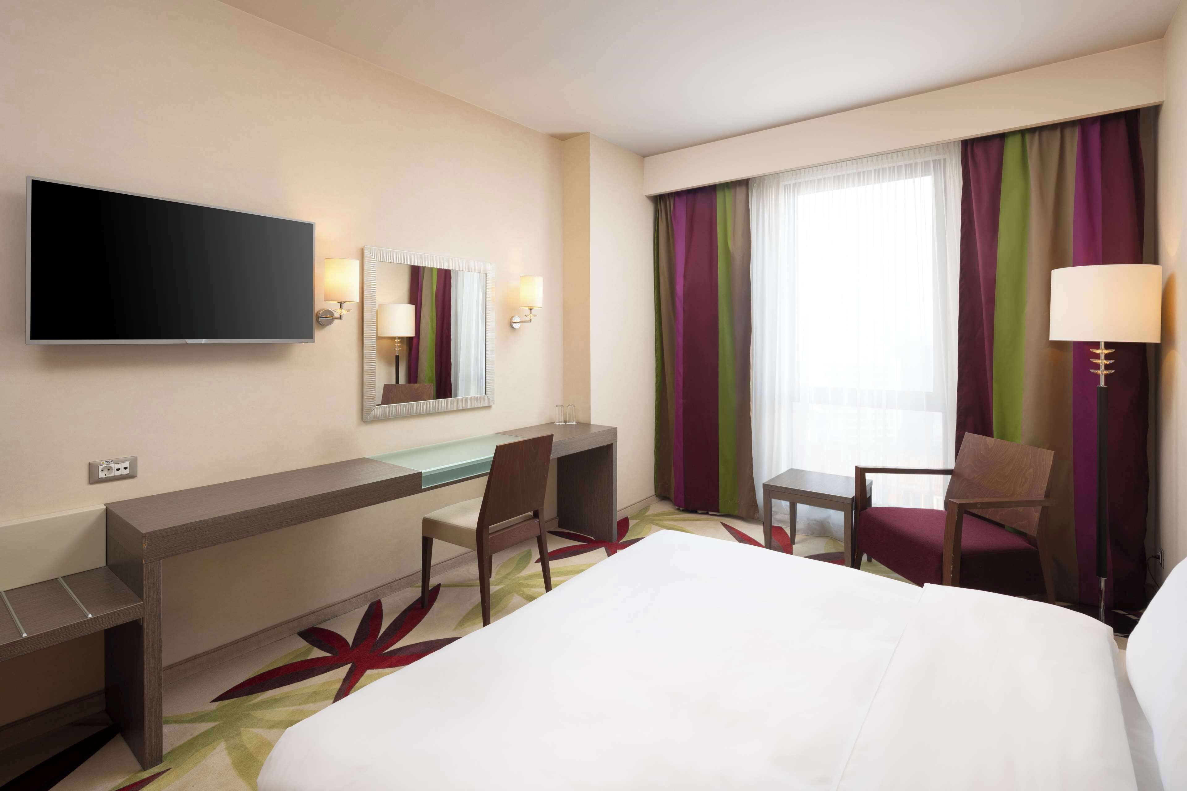 A hotel room with a bed, desk, chair, TV, and a window with curtains.