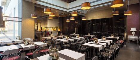 A spacious restaurant with multiple dining tables and chairs, large pendant lights, and a view of the city through large windows.