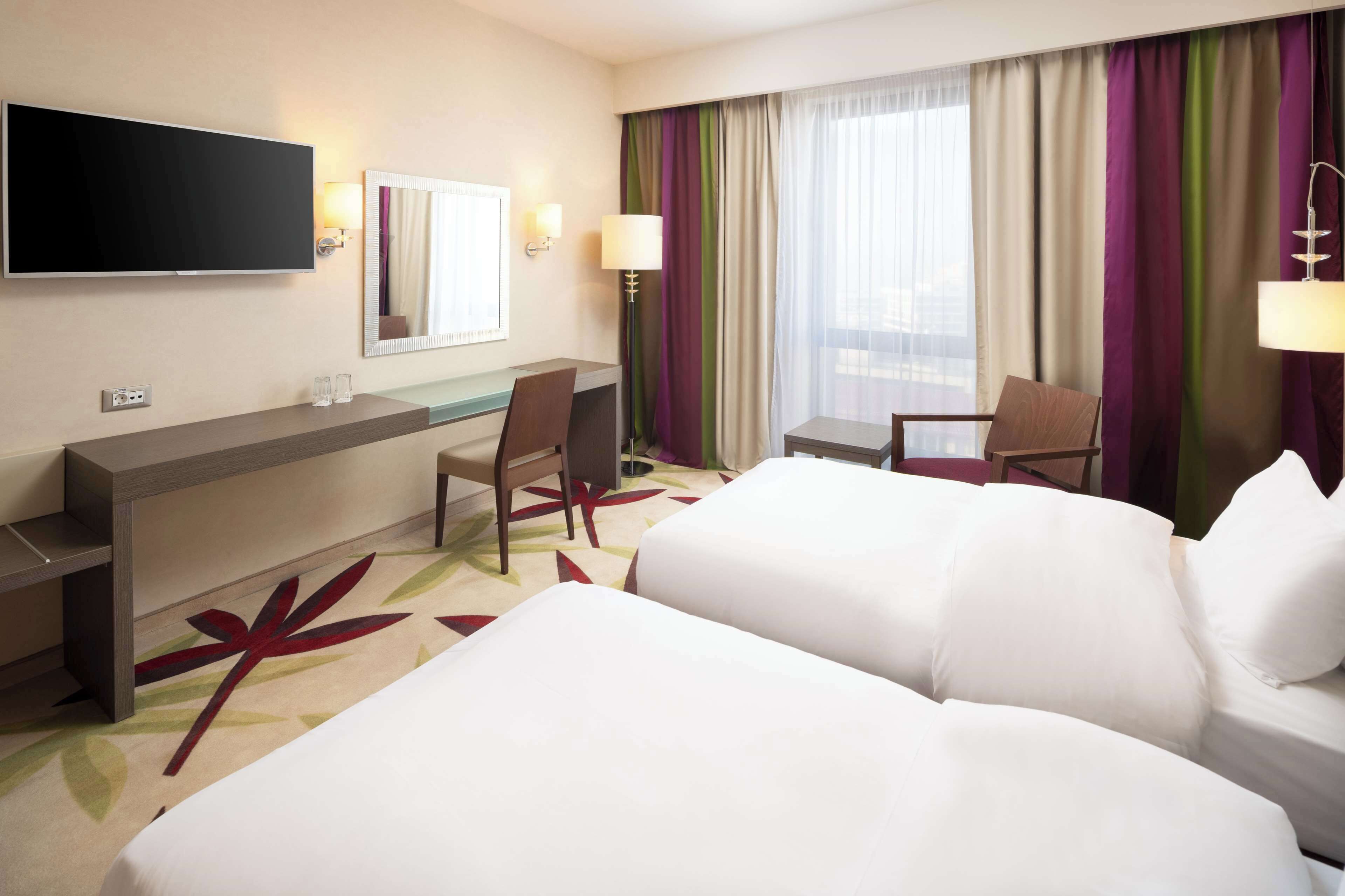 A hotel room with a bed, a desk, a chair, a TV, and a window with curtains.