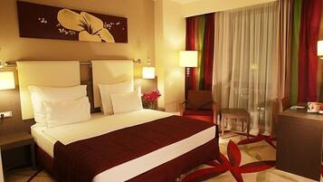 Habitación de hotel con una cama grande, dos mesitas de noche con lámparas, un escritorio con silla y un ventanal con cortinas.