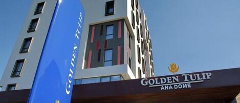 The exterior of the Golden Tulip ANA Dome hotel with a prominent blue flag and a modern architectural design.