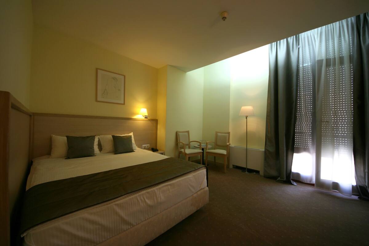 Une chambre d’hôtel avec un grand lit, deux chaises, une table et une fenêtre avec des rideaux.