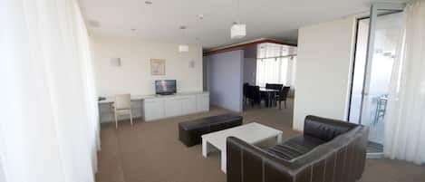 Una sala de estar moderna con un sofá, una mesa de centro y un televisor. Hay un comedor con una mesa y sillas. La habitación tiene paredes blancas y piso alfombrado.