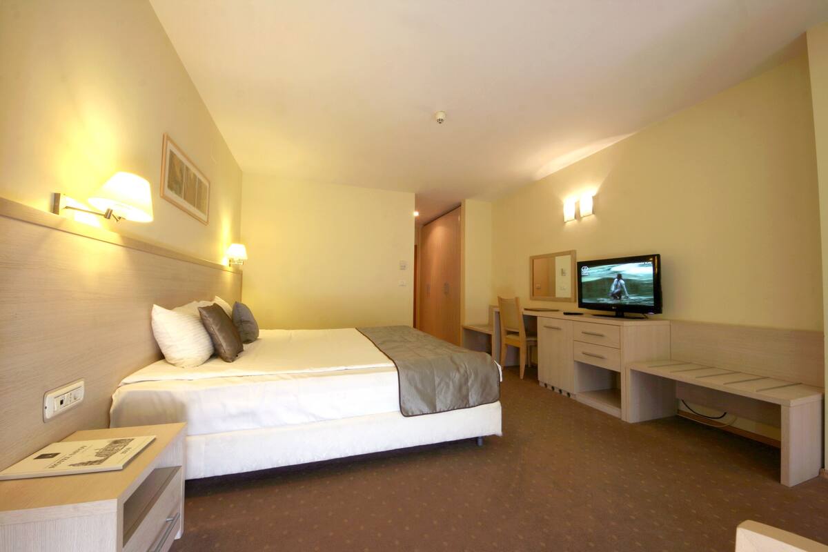 Une chambre d’hôtel équipée d’un lit, d’une table de chevet, d’un bureau, d’une chaise, d’une télévision et d’un luminaire fixé au mur.