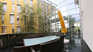 An outdoor dining area with a glass table and wicker chairs, surrounded by a yellow building and a glass wall. - Hotel Savoy (Timisoara)