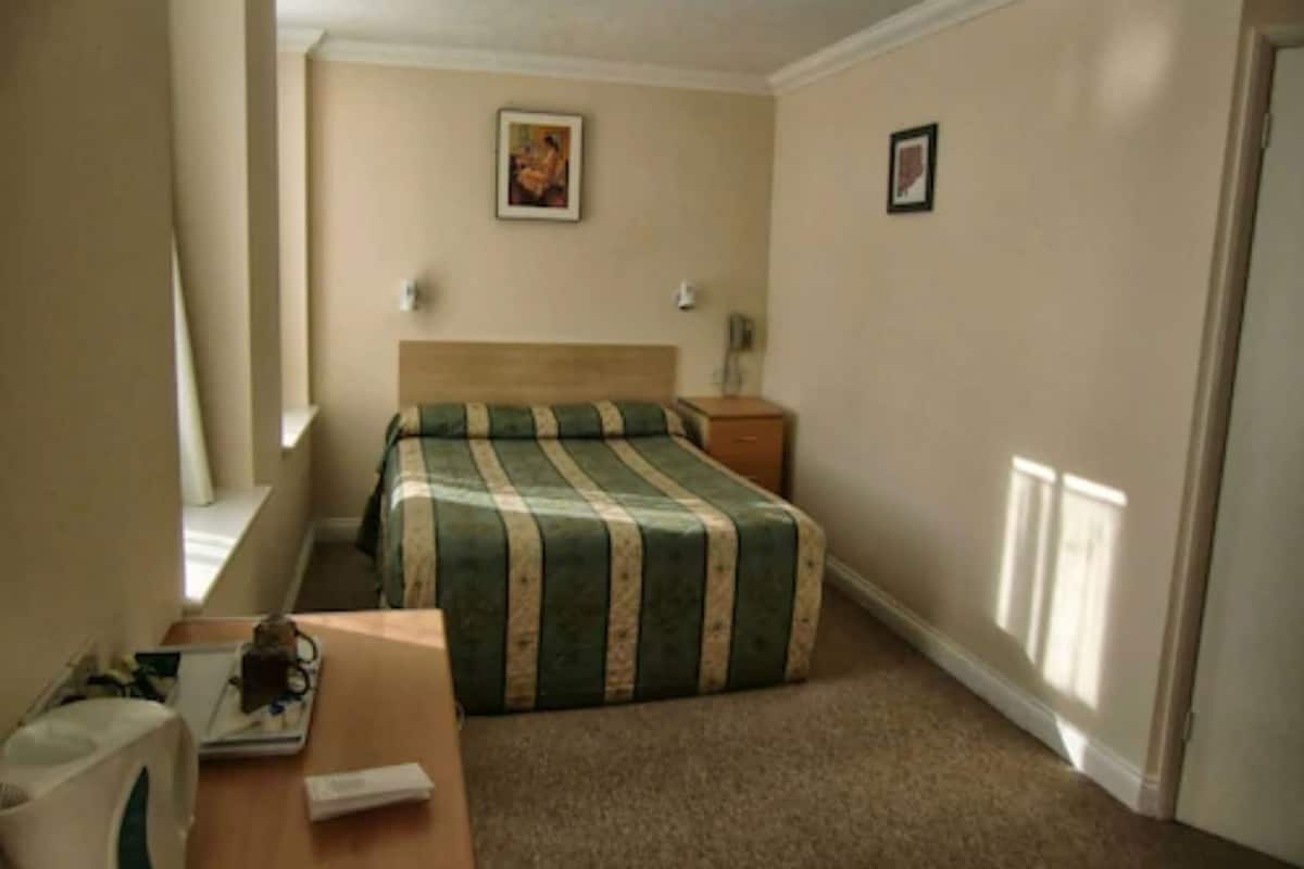 Une chambre d’hôtel avec un lit, une table de chevet, une lampe et une fenêtre avec des stores.