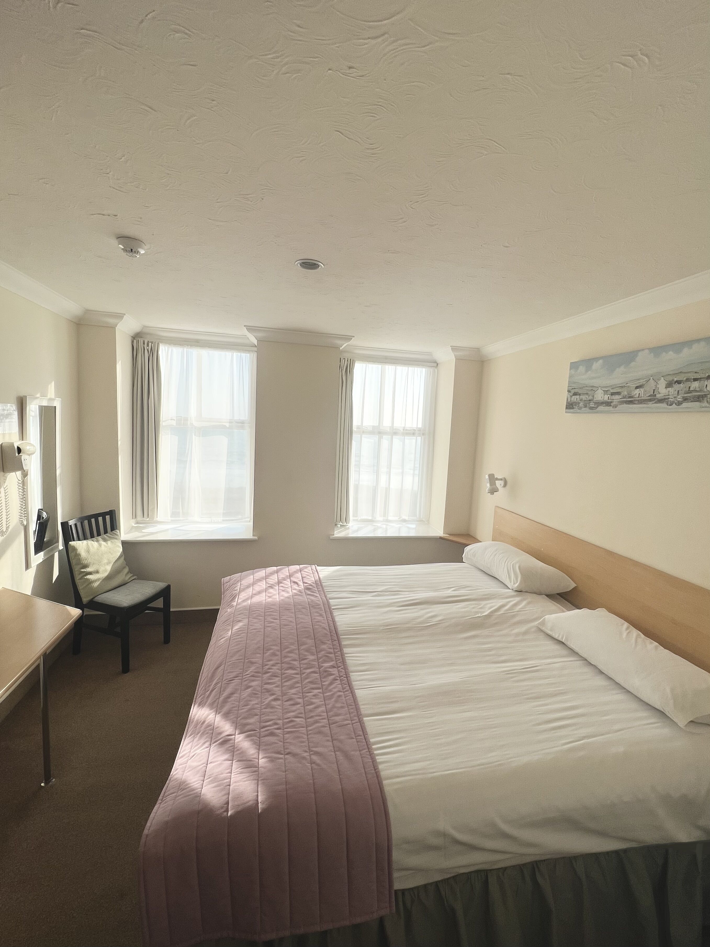 A hotel room with a bed, a chair, a desk, and two windows.