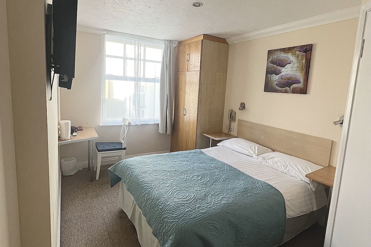 Une chambre d’hôtel avec un lit, un bureau, une chaise, un tableau au mur et une fenêtre avec des rideaux.