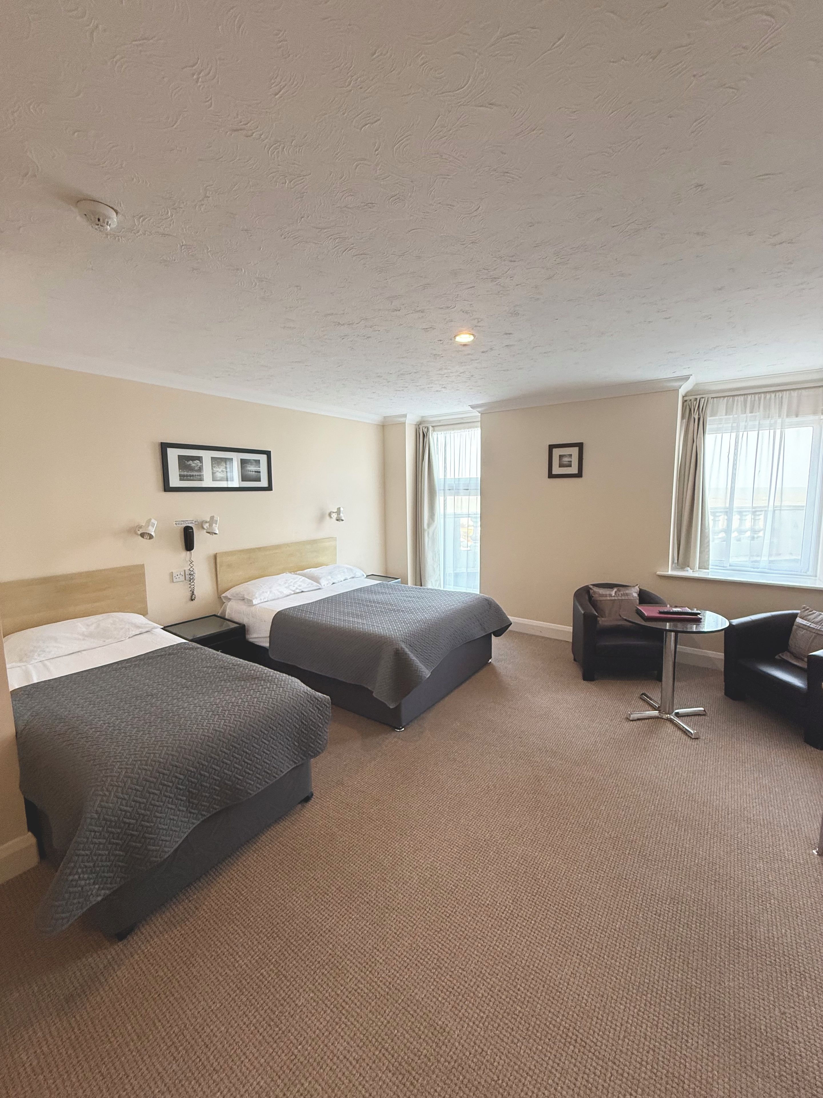A hotel room with two beds, a small table, a chair, and a window with curtains.