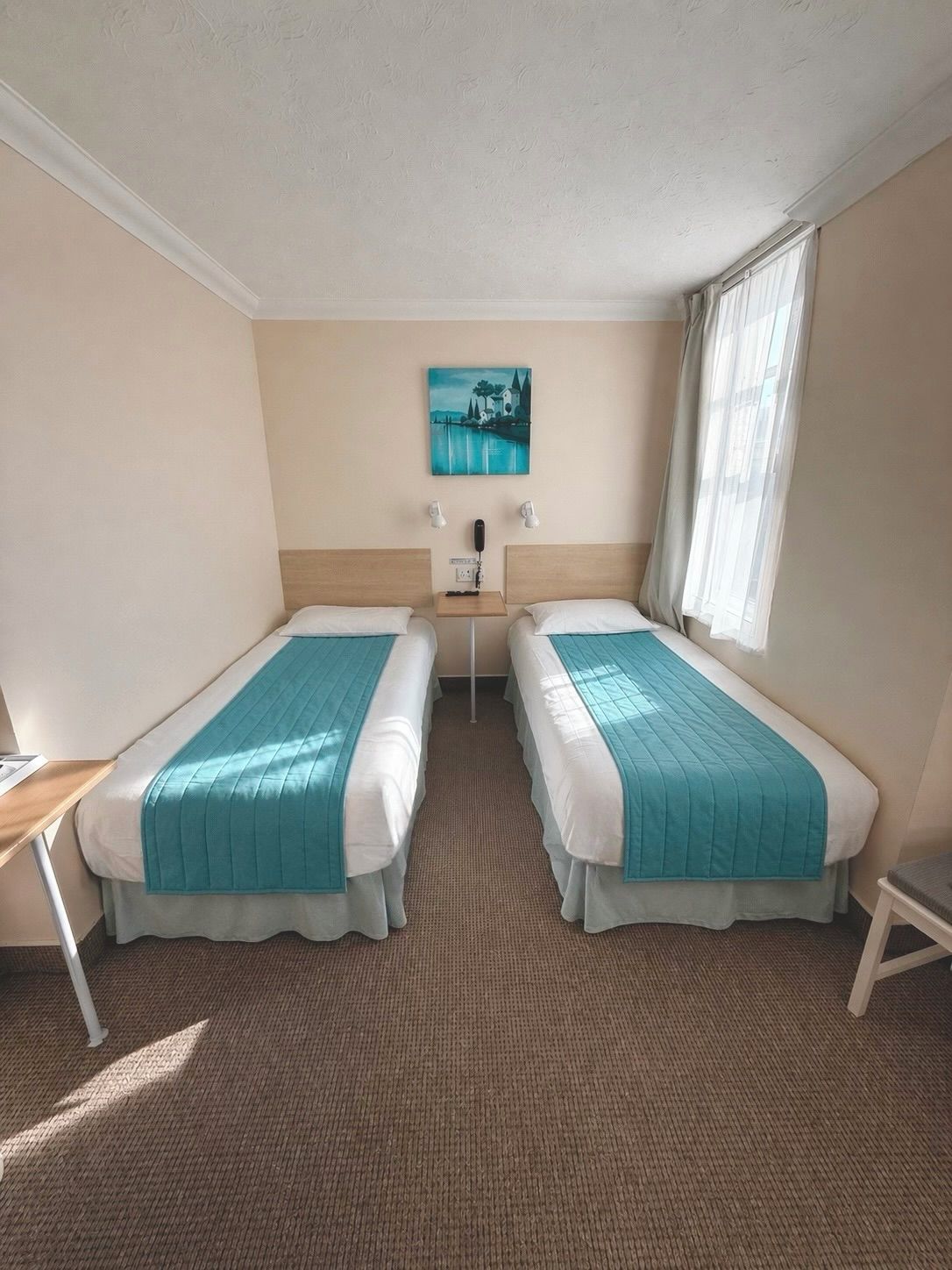 A hotel room with two beds, a small table, a painting on the wall, and a window with curtains.