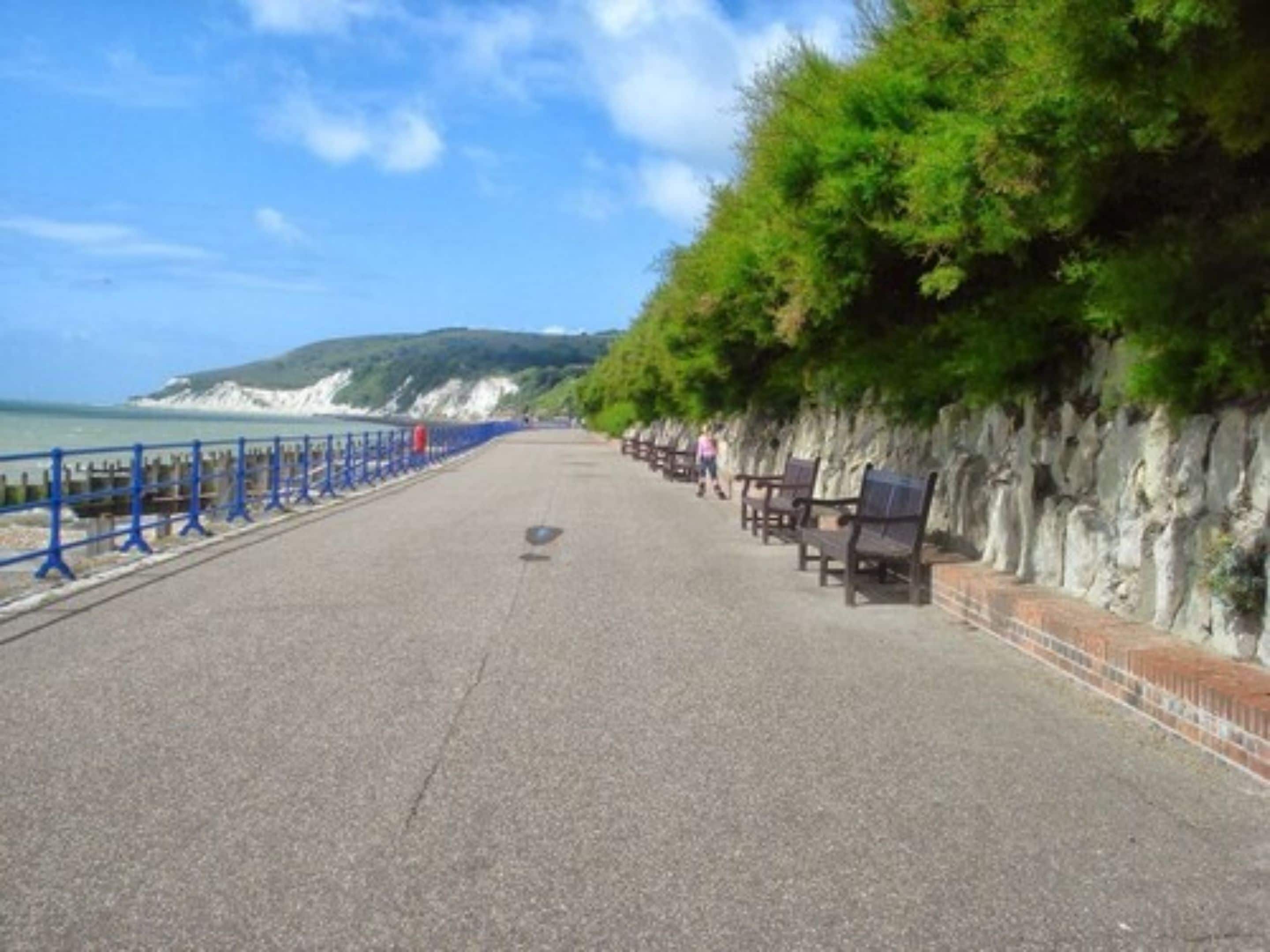 Un chemin pavé bordé de bancs, une rambarde bleue et une falaise rocheuse.