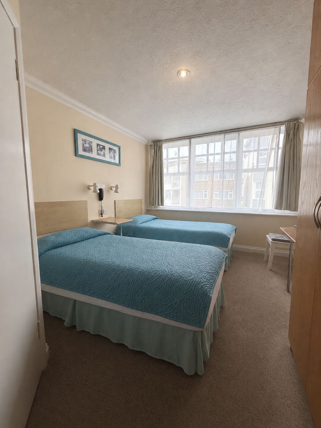 A hotel room with two beds, a bedside table, a chair, a window with curtains, and a wall-mounted phone.
