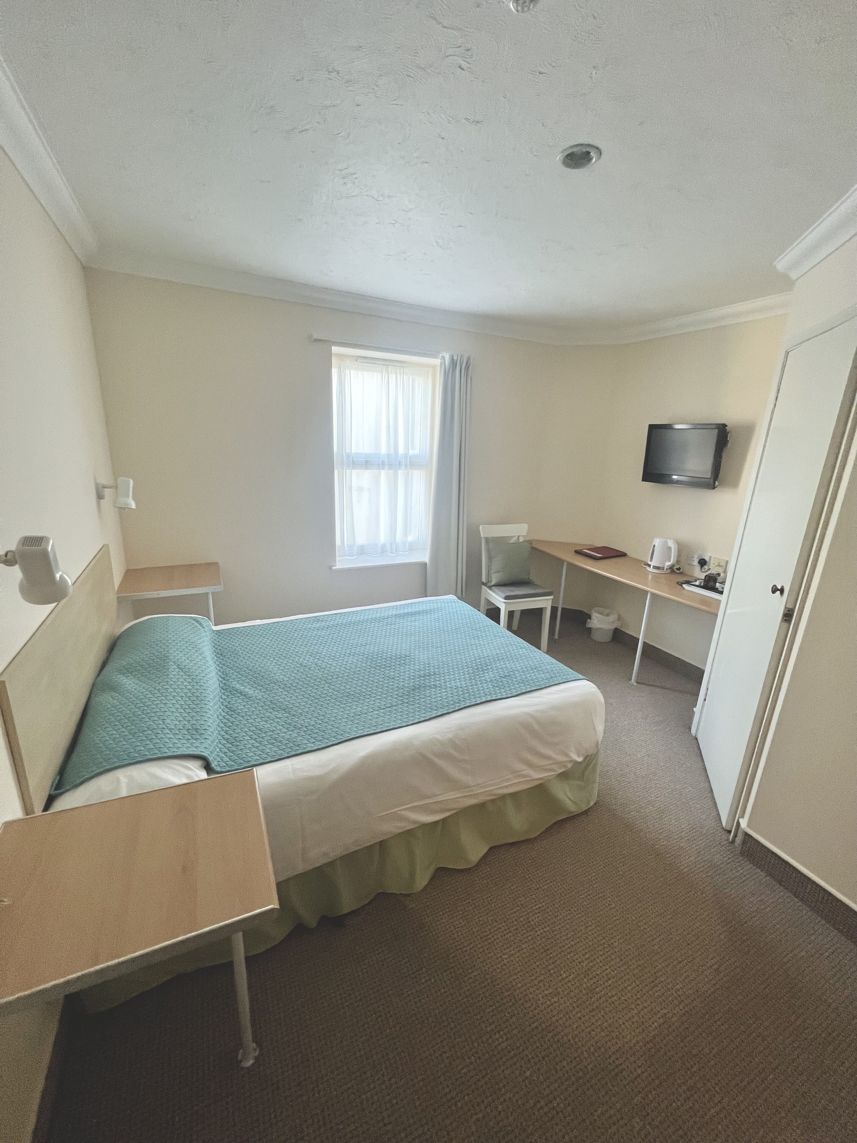 A small, neatly arranged hotel room with a bed, a desk, a chair, a television, and a window with curtains.