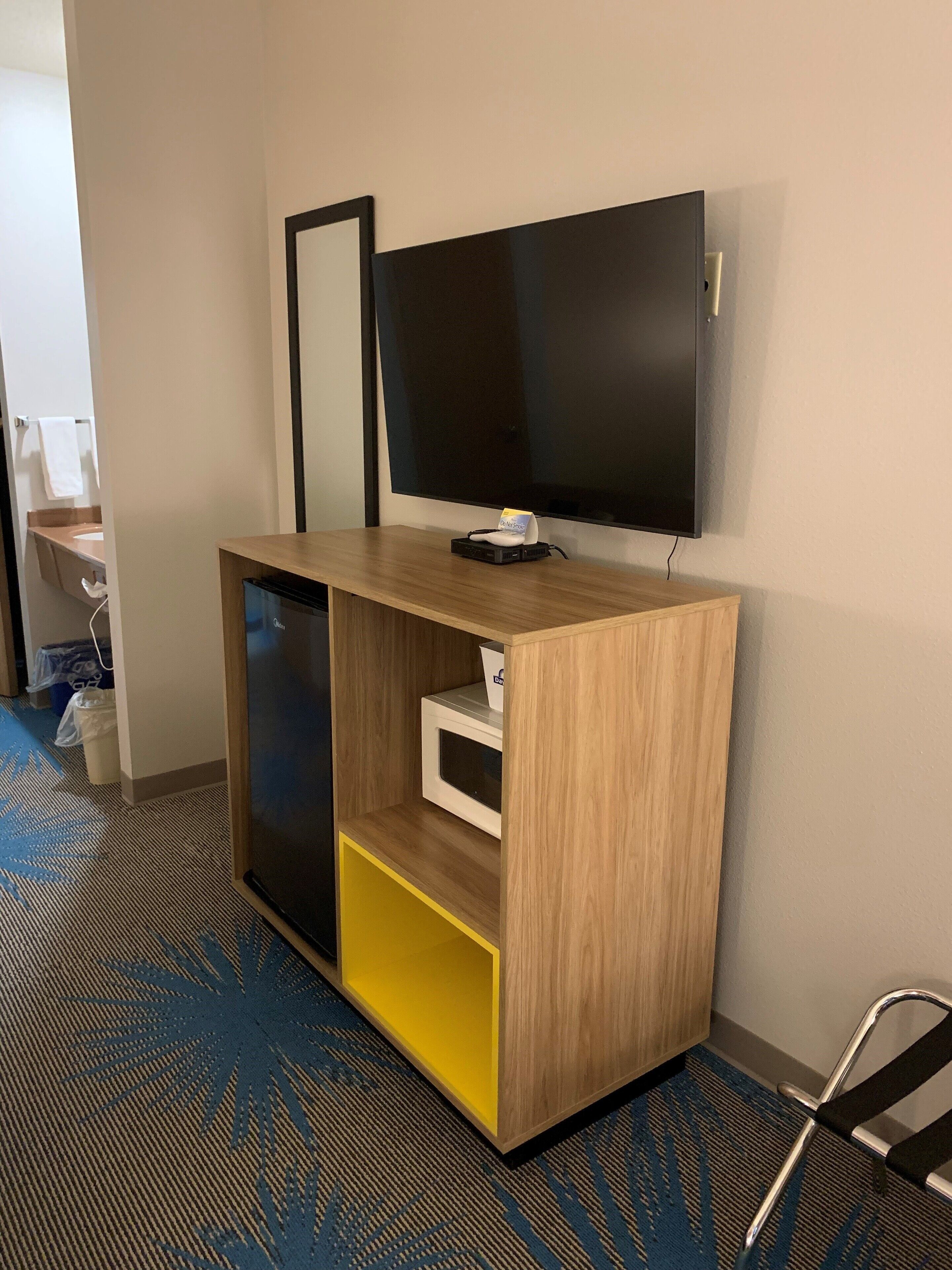 A hotel room with a wooden desk, a flat-screen TV mounted on the wall, a microwave, and a small cabinet.