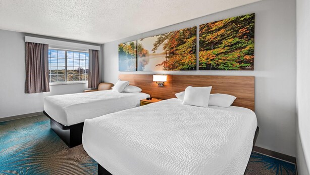 Una habitación de hotel con dos camas, un ventanal con cortinas, un cuadro en la pared y una mesita de noche con lámpara.