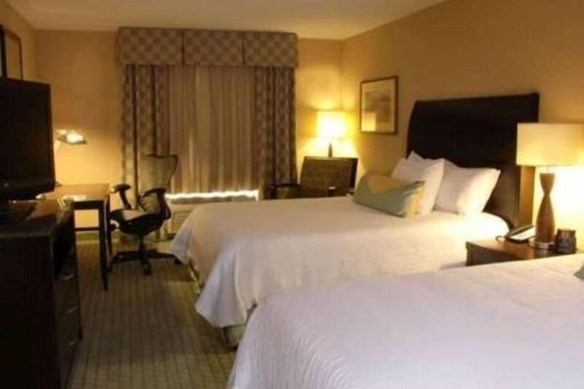 Ein Hotelzimmer mit zwei Betten, einem Schreibtisch, einem Stuhl, einem Fernseher und einem Vorhangfenster.