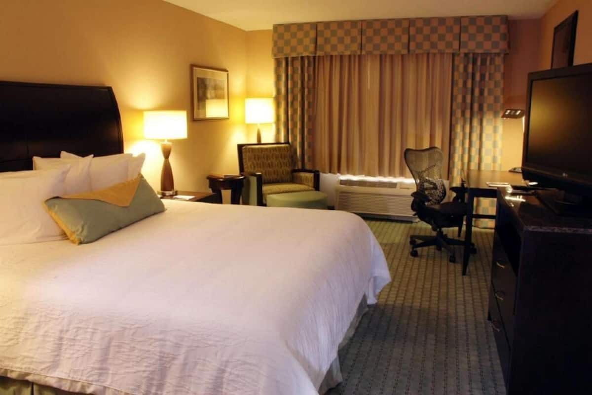 Ein Hotelzimmer mit einem großen Bett, einem Schreibtisch, einem Stuhl, einem Fernseher und einem Vorhangfenster.