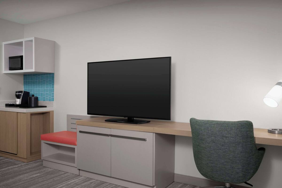 A modern room with a flat-screen TV mounted on a wall, a desk with a chair, a small kitchenette, and a tiled backsplash.
