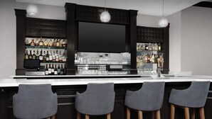 Una zona de bar con mostrador blanco, taburetes de madera oscura y estantes llenos de botellas y vasos.