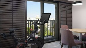 Una habitación moderna con un amplio ventanal corredero que da a un balcón, una bicicleta estática, una mesa redonda y una sola silla.