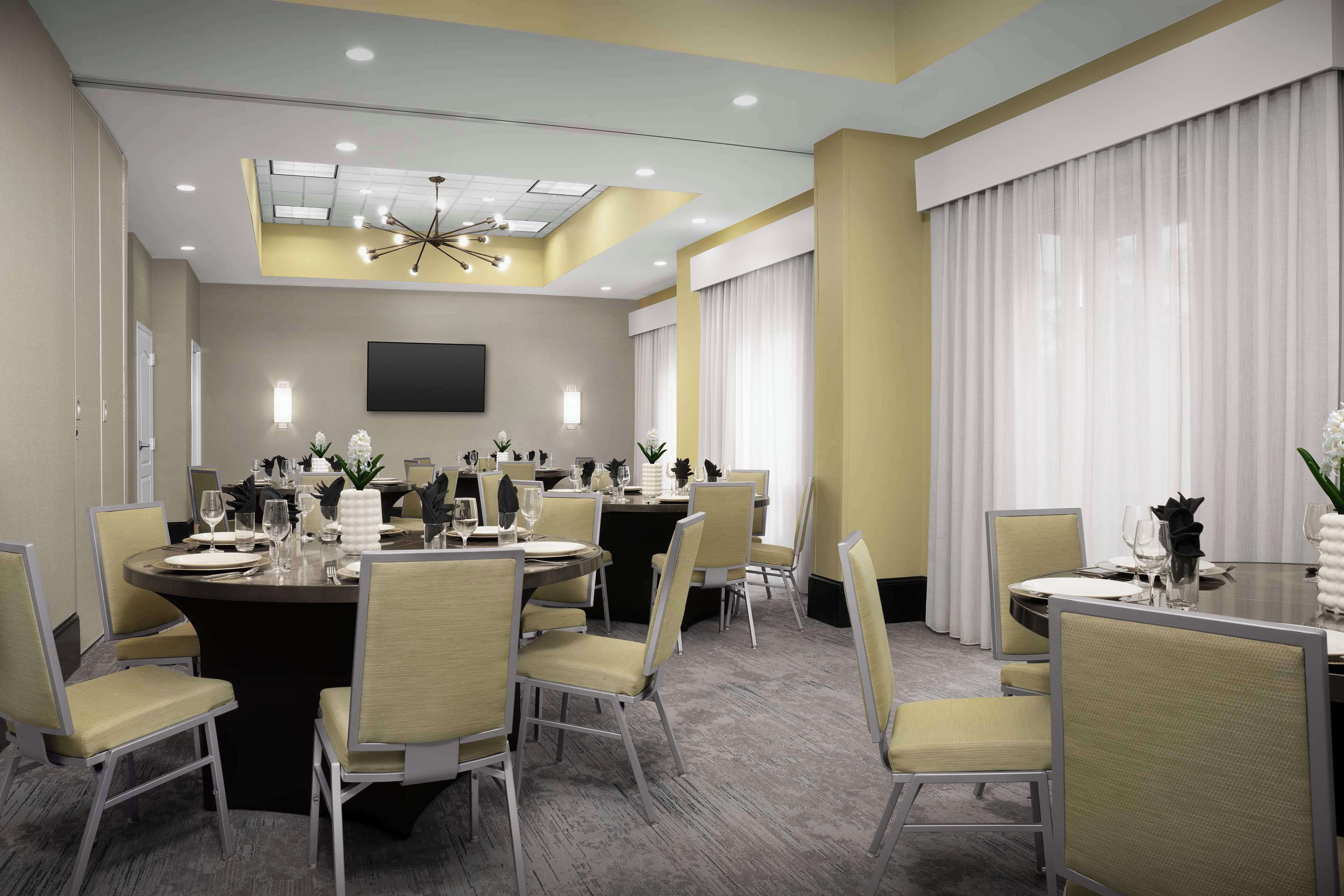 A conference room with a large round table, multiple chairs, a flat-screen TV, and a ceiling light fixture.
