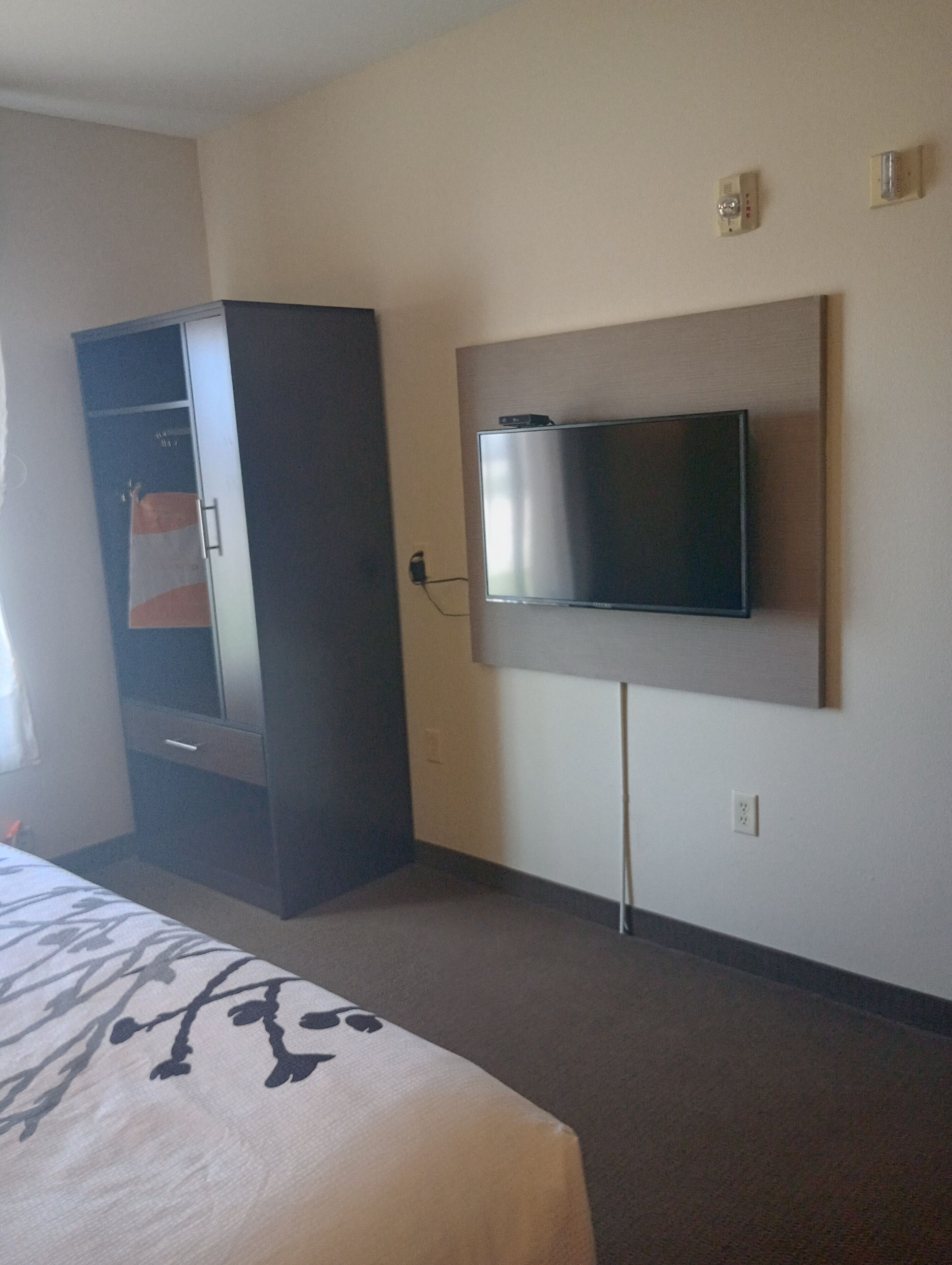 A hotel room with a bed, a wardrobe, a television mounted on the wall, and a power outlet.