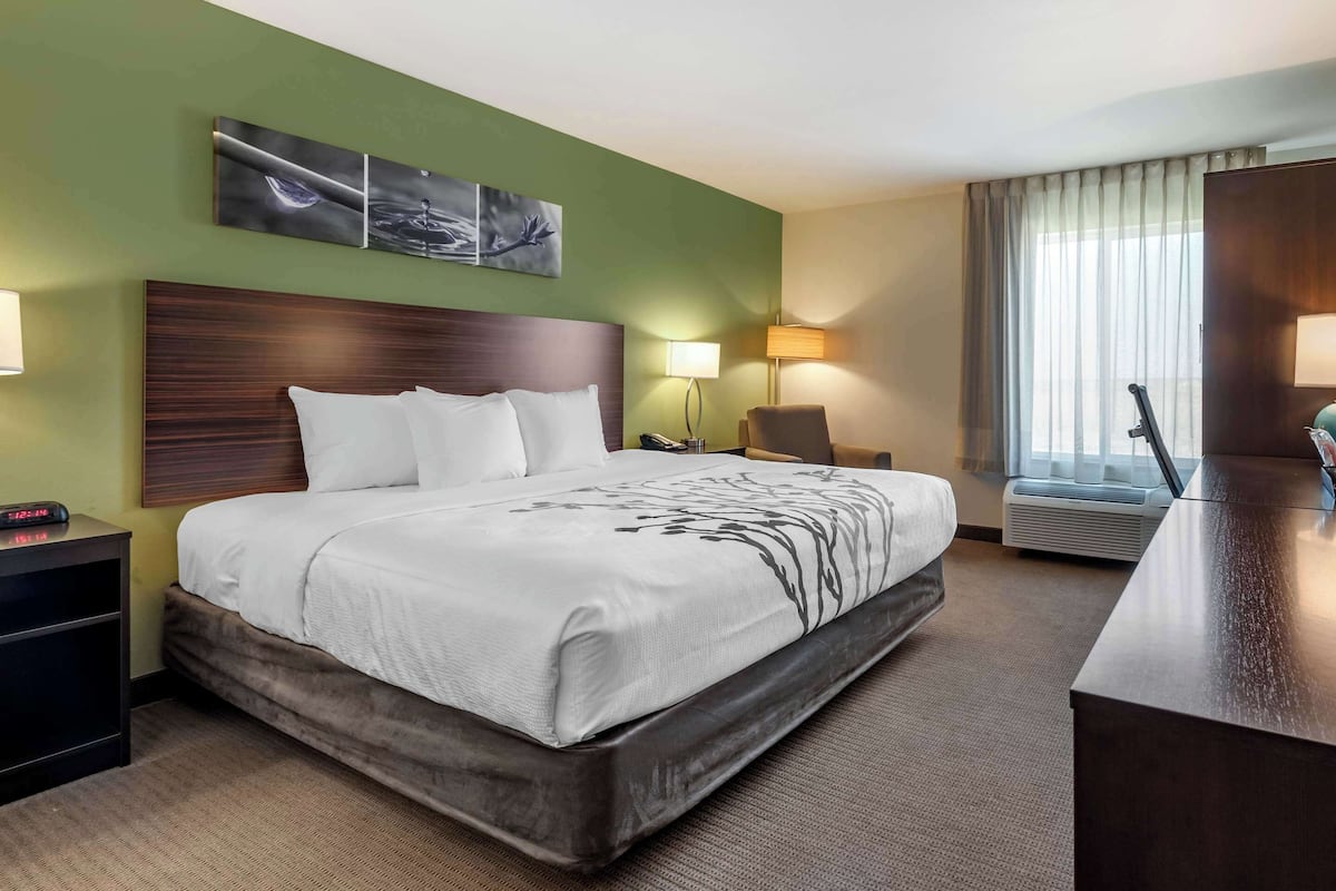 A hotel room with a large bed, a wooden headboard, a desk with a chair, a lamp, and a window with curtains.