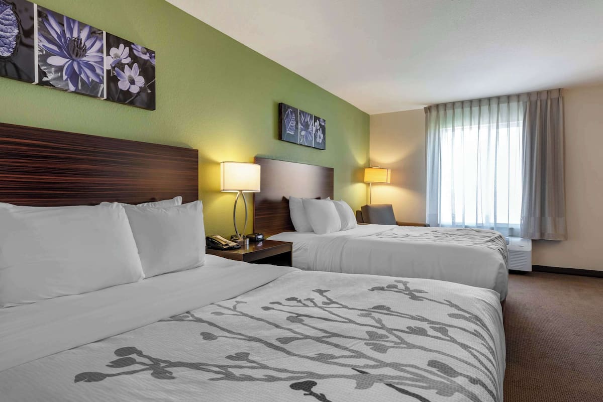 A hotel room with two beds, a wooden headboard, a nightstand with a phone, two wall-mounted lamps, and a window with curtains.