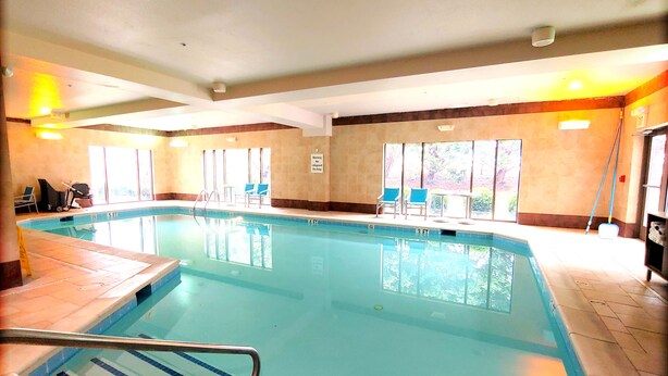 An indoor swimming pool with clear water, a diving board, and a sunlit area with chairs and a table.