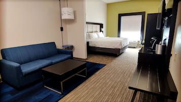 Quarto de hotel com sofá azul, cama com roupa de cama branca, uma mesinha, televisão e secretária com luminária.