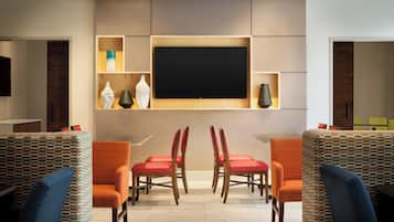 A modern interior with a flat-screen TV mounted on a wall, surrounded by shelves with decorative items. There are two wooden chairs with red cushions and a patterned sofa.