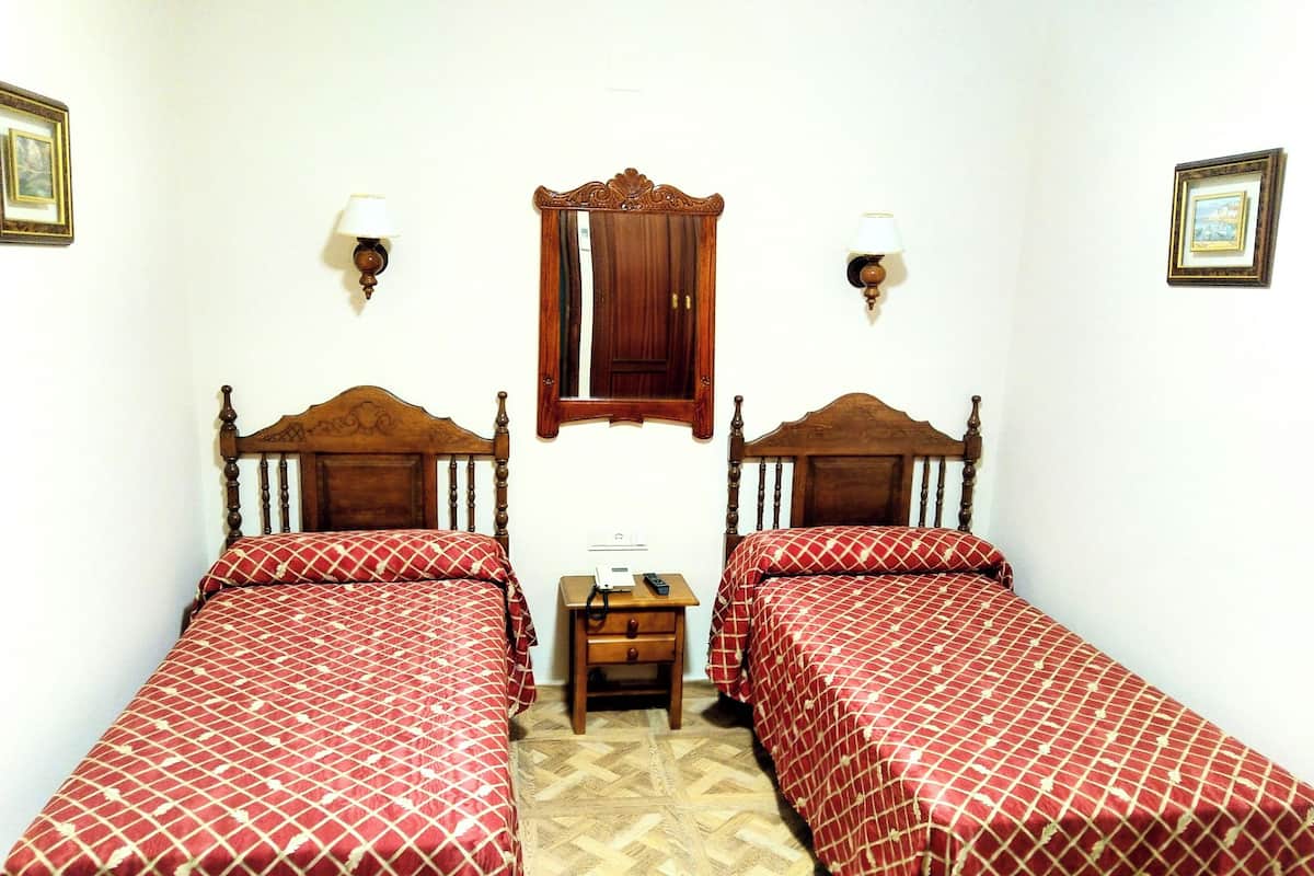 Une chambre avec deux lits en bois, une petite table de chevet en bois, un miroir et deux lampes fixées au mur.