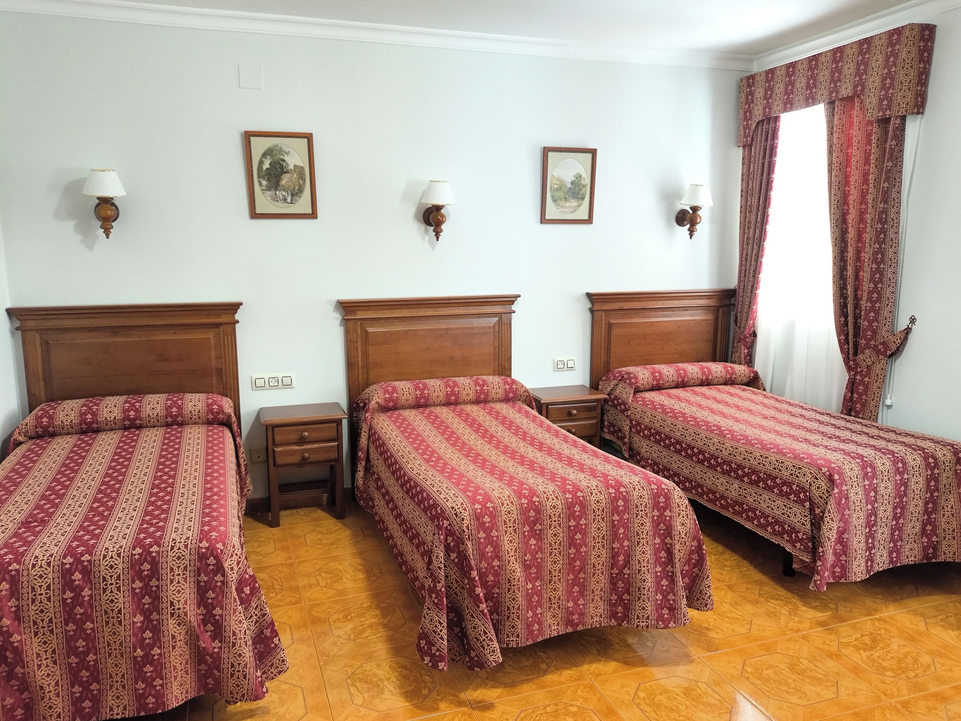 A hotel room with three wooden beds, each with a patterned bedspread. There are two wall-mounted lamps and two framed pictures on the wall.