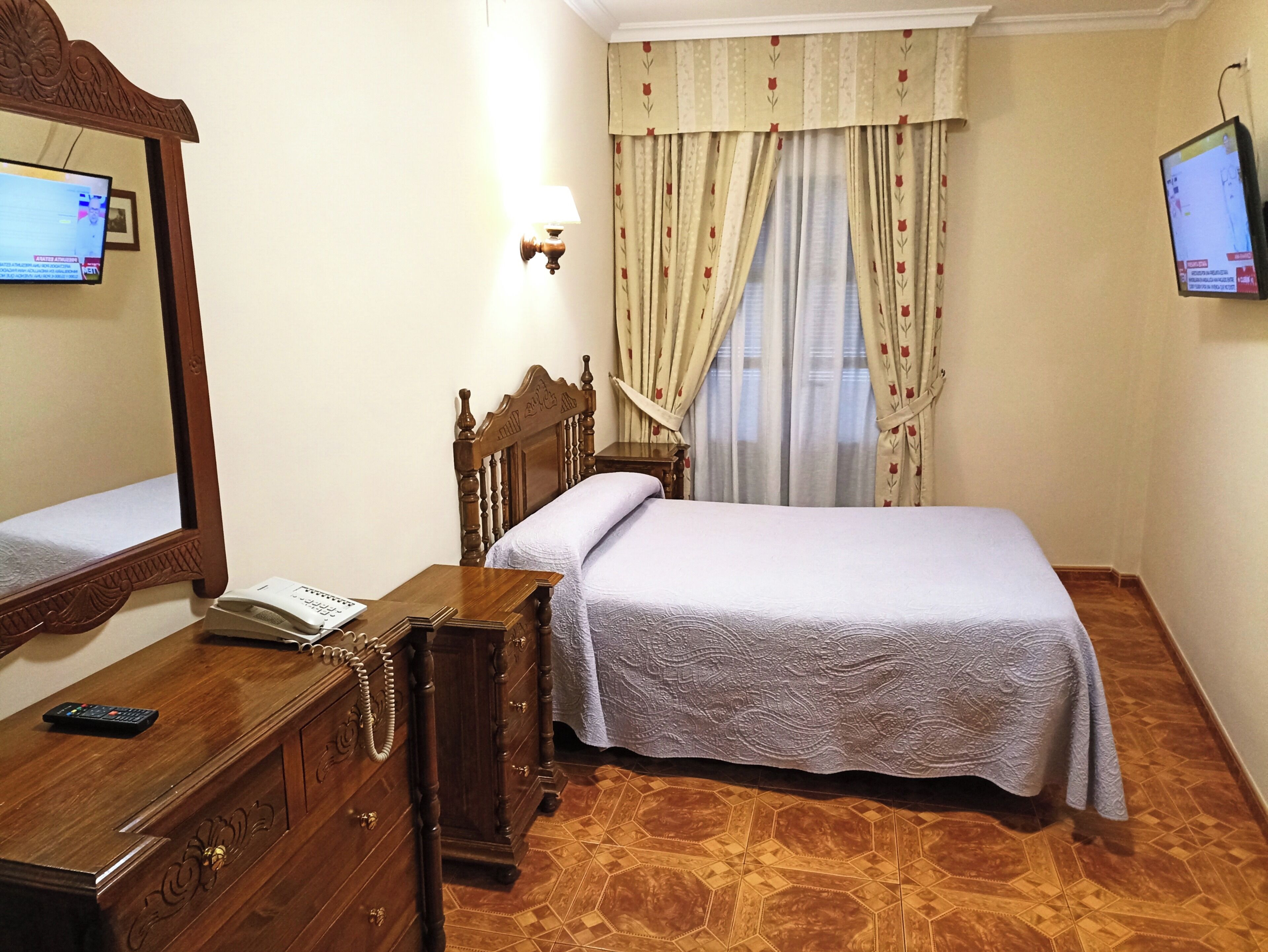 A hotel room with a wooden bed, a nightstand with a telephone, a mirror, and a television on the wall.