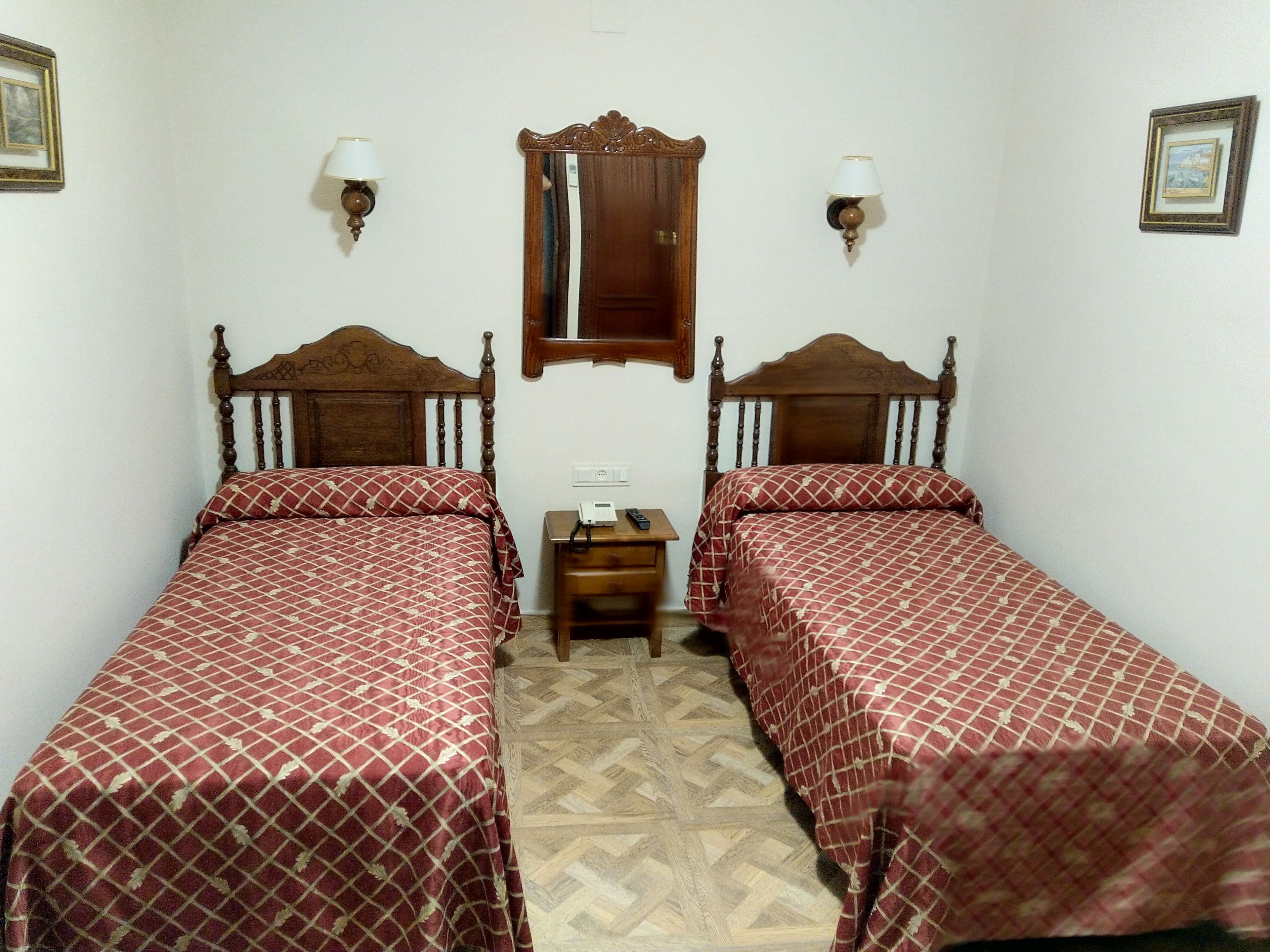 A room with two wooden beds, a small bedside table, a mirror, and wall-mounted lamps.