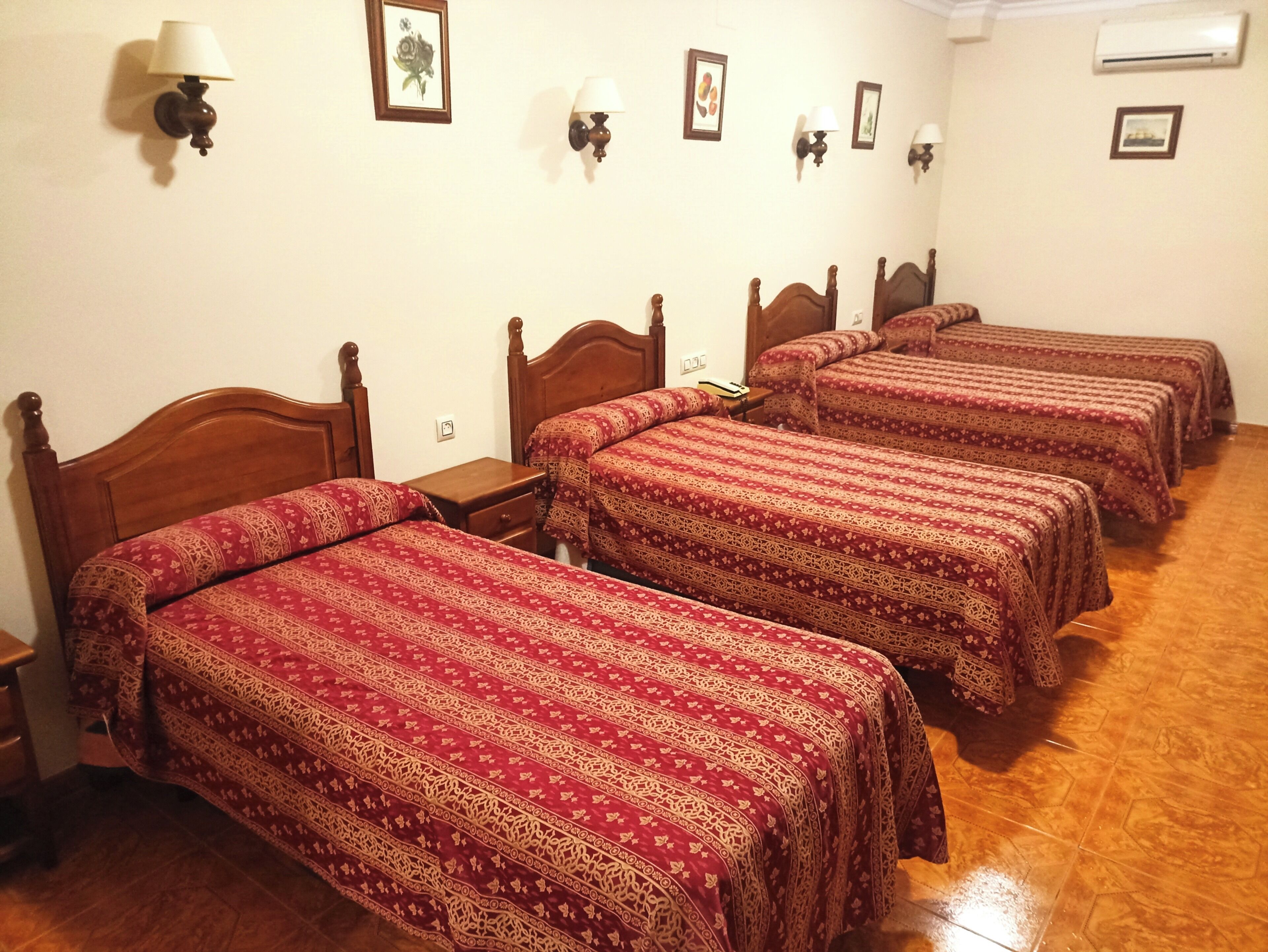 A hotel room with four wooden beds, each with a red patterned bedspread. There are wall-mounted lamps and framed pictures on the walls.