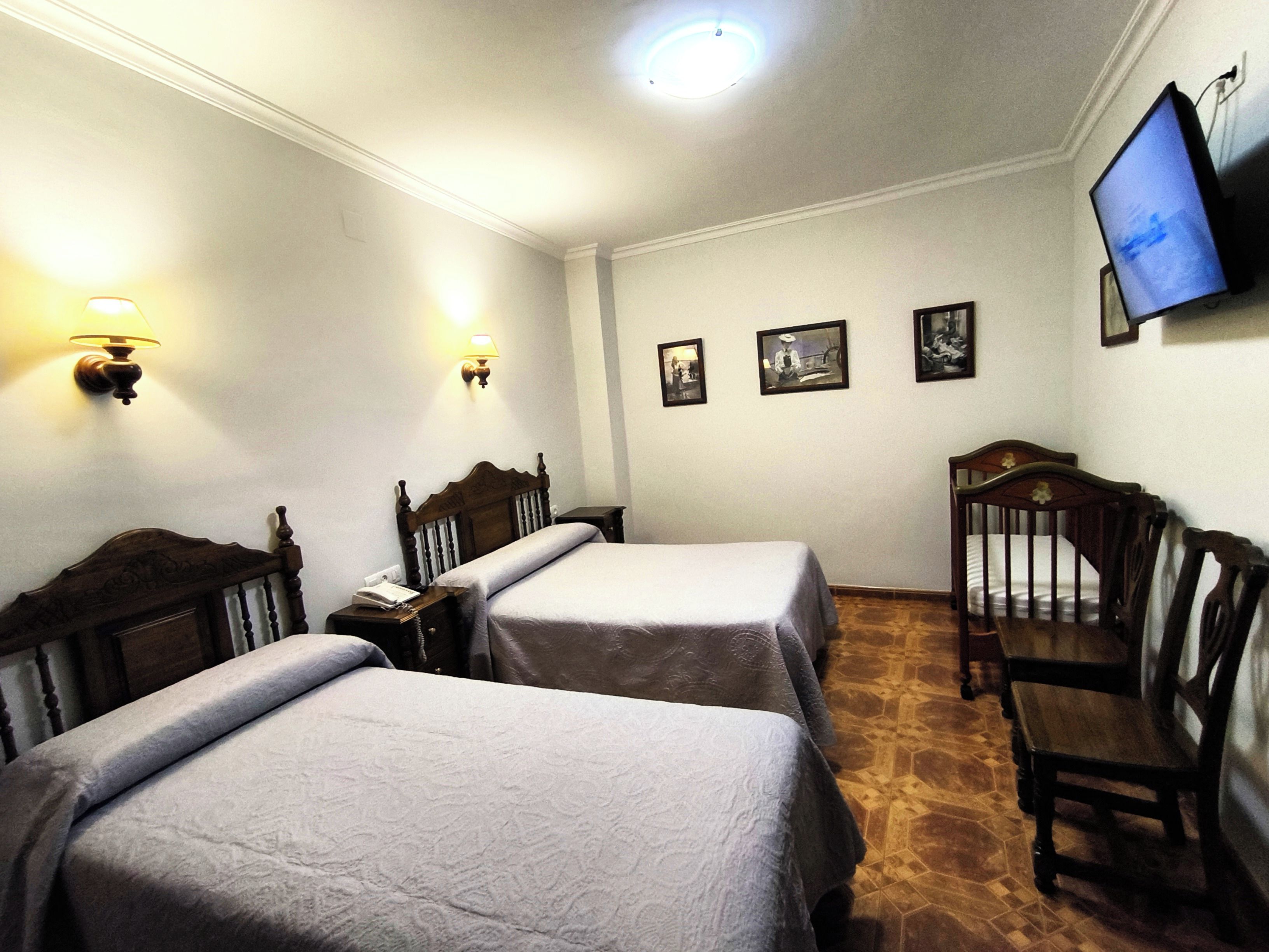 Habitación de hotel con dos camas individuales, una silla de madera, un televisor fijado a la pared y dos lámparas de pared.