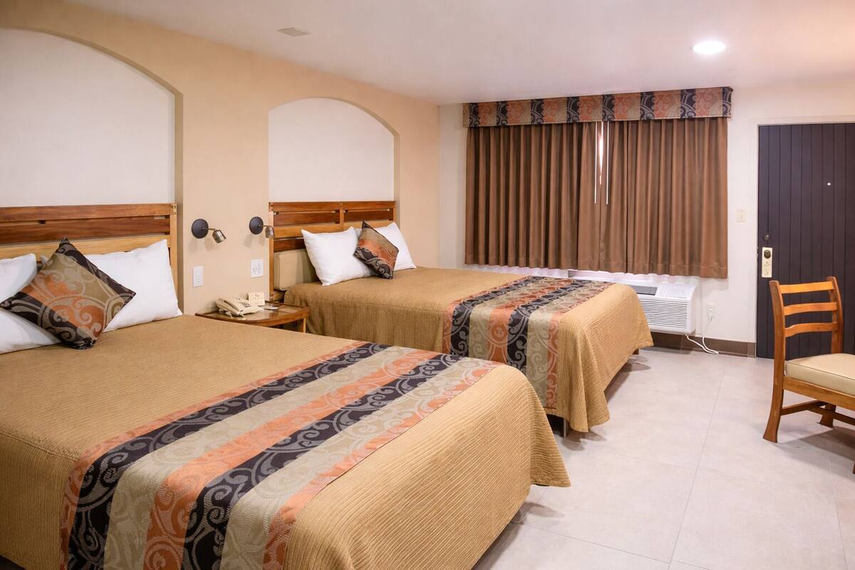 Ein Hotelzimmer mit zwei Betten, einem Holzsessel, einem Nachttisch mit Telefon und einem Fenster mit Vorhängen.
