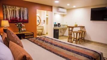 Quarto de hotel com cama, escrivaninha, cadeira, cozinha compacta e televisão.