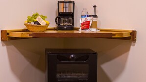Une petite cuisine équipée d’une machine à café, d’un panier de fruits et d’un distributeur de capsules de café, le tout placé sur une étagère en bois.